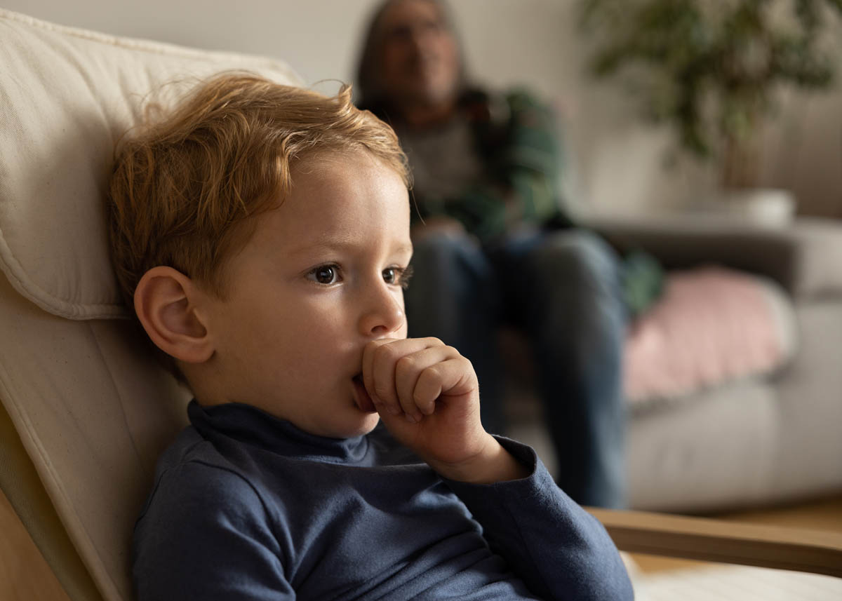 A little boy sits at home in a chair and sucks his finger, in the background a man watches the boy. High quality photo