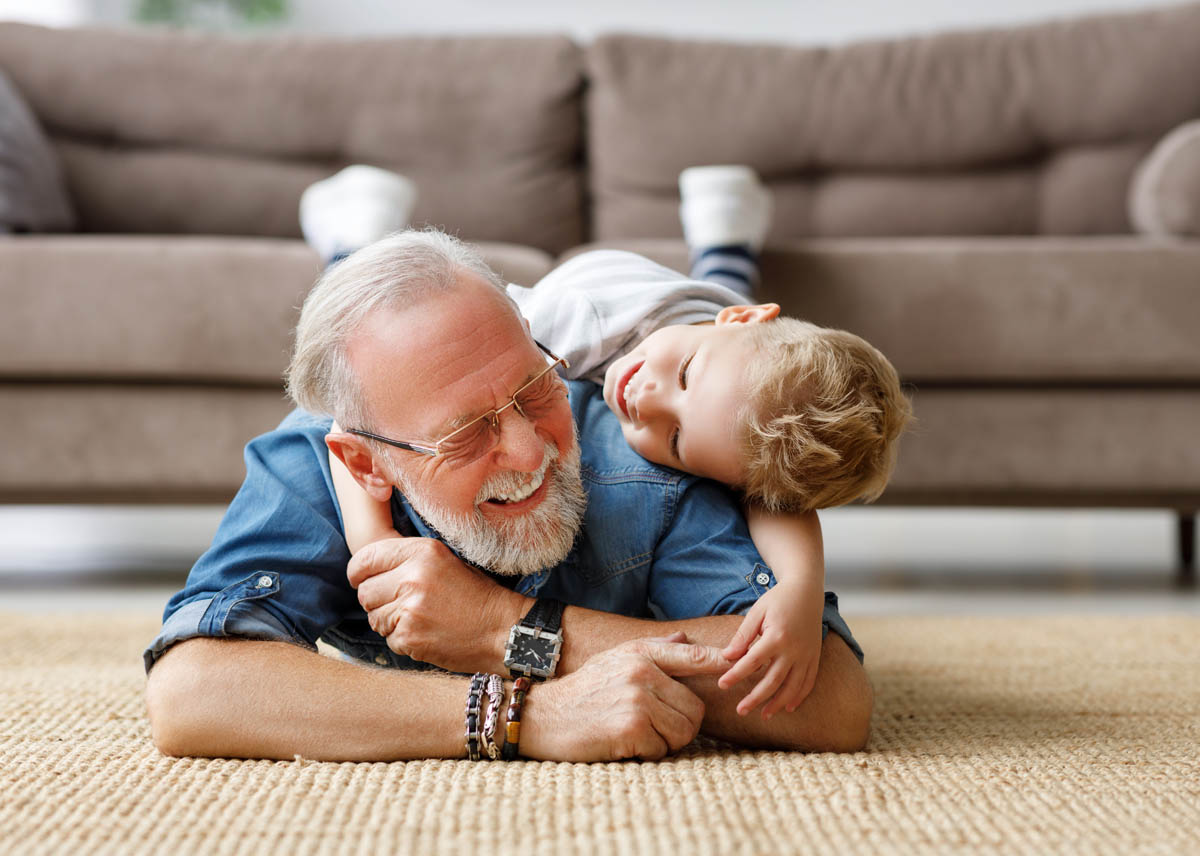 Happy family: delighted boy  embracing elderly grandfather from behind and looking at camera while lying on floor  on weekend day at home